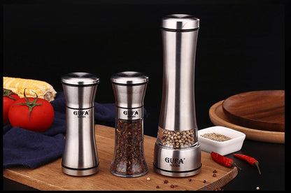two small pepper and salt stainless steel grinders besides a big stainless grinder on a wood support decorated with food around
